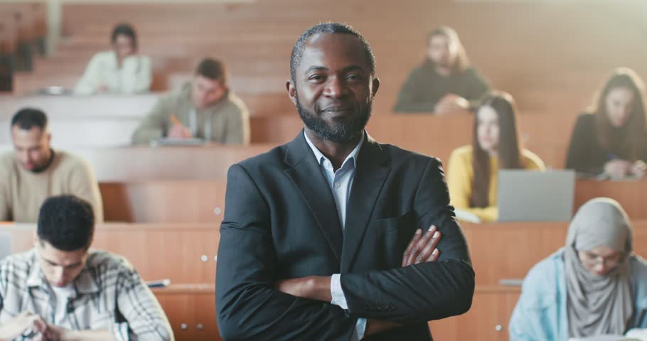 Portrait shot of handsome happy African American male professor crossing hands at University, looking at camera and laughing. Students in class on background. Man teacher of high school. Study concept | Shutterstock HD Video #1101012837