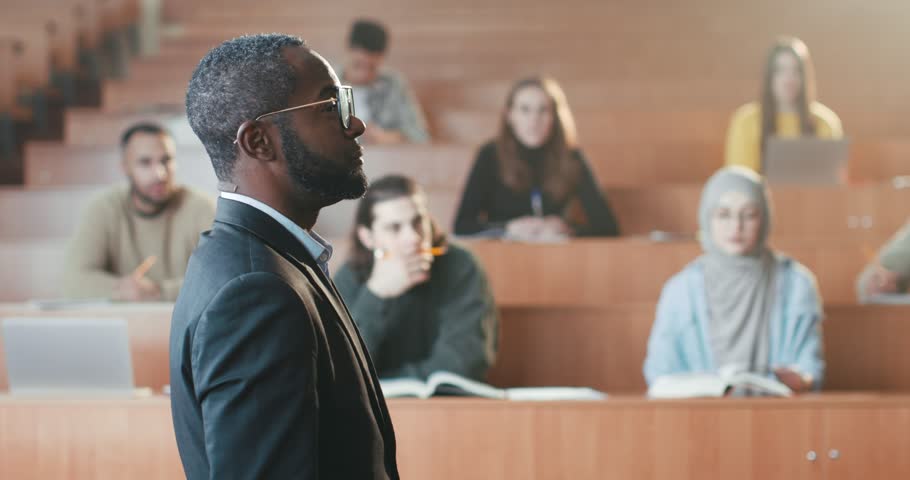 Rear of African American male professor standing in front of students in college and talking to them while having lection. High school. Back view. Man teacher explaining subject at lesson. Close up. Royalty-Free Stock Footage #1101012861