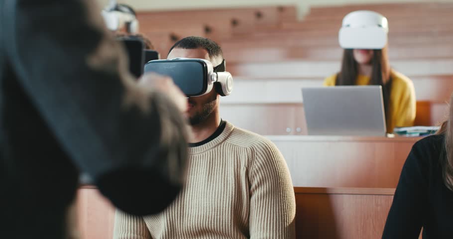 Back view of professor talking with laptop in front of mixed-races students in VR glasses. Multiethnic males and females learning high tech and listening to teacher at lection. Techonology education.