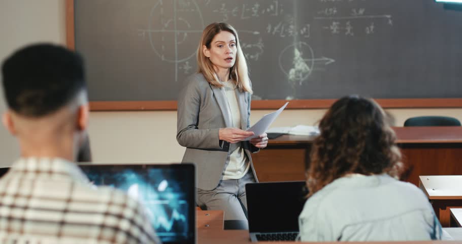 Beautiful Caucasian female professor explaining lection at University in front of students sitting at desks. Education concept. Woman teacher speaking to young adults at college. Geometry math lesson. | Shutterstock HD Video #1101012985