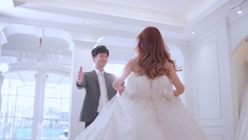 Asian beautiful bride trying on wedding gown with her fiance in studio. Attractive young couple feeling happy and relax, enjoying preparation for marriage ceremony in fitting room at the bridal store.