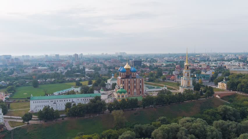 Ryazan, Russia. The Ryazan Kremlin, the oldest part of the city, is seen from above with the Cathedral of the Assumption of the Blessed Virgin Mary as its dominant centerpiece. The cathedral, built between 1684 and 1699 by architect Yakov Bukhvostov after an earlier structure collapsed, rises over 40 meters with five large drums and cupolas in blue and gold, and is larger than the Dormition Cathedral in the Moscow Kremlin. The surrounding Kremlin complex includes white-walled historic buildings with green roofs, a tall neoclassical bell tower, and extensive parkland. The scene is set on a bright summer day with soft daylight, revealing the contrast between the red brick cathedral, white stone ornamental trim, and the dense green canopy of trees that frames the ancient citadel against the modern city skyline in the distance. The slow aerial orbit showcases the full scale and architectural harmony of this historic Russian Orthodox landmark, Point of interest
