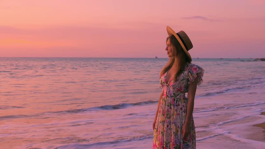 Female tourist in dress stands on the beach at a pink sunset, looking at the camera. Happiness of freedom on summer vacation by ocean. Woman enjoying tropical holidays. Slow motion. Royalty-Free Stock Footage #1101024609
