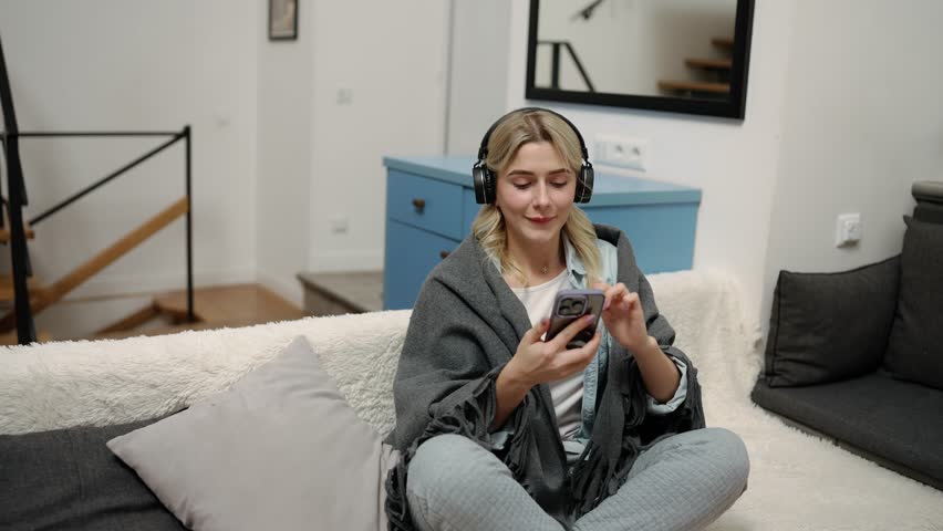 A thirty-year-old woman sits on a bed in headphones and listens to music | Shutterstock HD Video #1101025665
