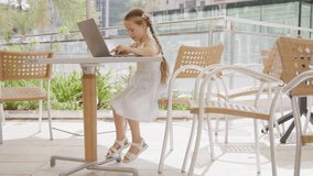 Portrait of cute, caucasian girl with blonde hair smiling while having a video call outdoors. Smart kid using a laptop to play funny games and watching cartoons. High quality 4k footage