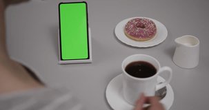 Woman Sitting at Table Coffee Donut Using Smartphone With Chroma Key Green Screen, Scrolling Through Social Network Media Online Shop Internet. Smartphone in Horizontal Mode with Green Screen Mock-up.