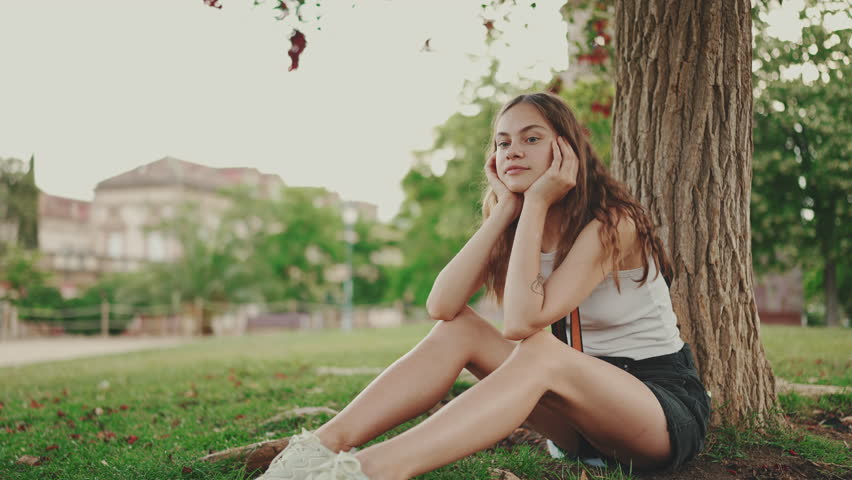 Girl is resting in the park. Beautiful girl looks around sitting on the lawn under tree in the park
