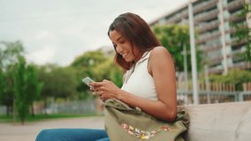 Beautiful girl with long dark hair wearing white top sits on bench and uses mobile phone. Young smiling woman writing in social networks on cellphone. Sideways movement