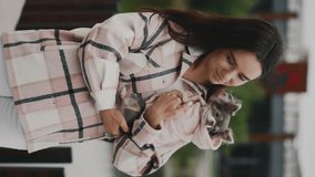 Vertical Video of the Beautiful Caucasian Woman holding the Chihuahua wearing Stylish Dog Clothes, standing looking at the camera on the blurred background. Portrait of Pretty Girl with her Pet