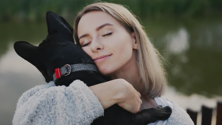 Happy Caucasian Woman Pet Owner with Closed Eyes Is Hugging and Kissing Her Black French Bulldog Puppy, Showing Love to the Dog Standing in the Park near the Local Lake. Close Up | Shutterstock HD Video #1101028521