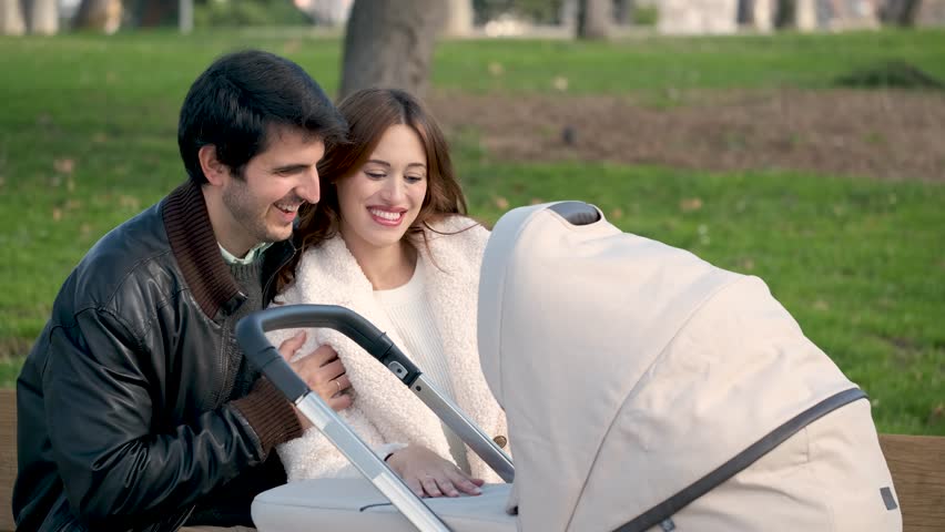 Couple talking to their baby in a stroller, kissing and hugging sitting on a bench in a park.