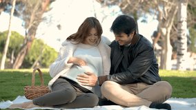 Man and pregnant woman looking at the belly on a picnic with their dog in a park. Family.