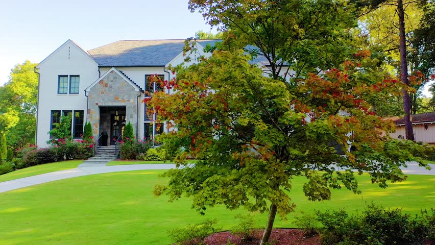 Drone shot slowly panning past a tree with fall colors to reveal a beautiful mansion with green grass in the front yard in Atlanta, Georgia.
