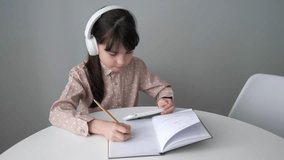 Cute child girl wearing headphones studying online on mobile phone. School kid holding smartphone doing homework at home using app and write in notebook. Digital education concept