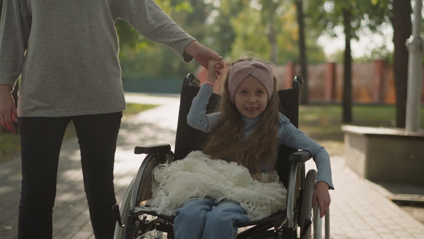 Toothless little girl smiles rotating wheel of medical equipment and holding hand of mom. Woman enjoys spring walk in park with daughter closeup | Shutterstock HD Video #1101038943