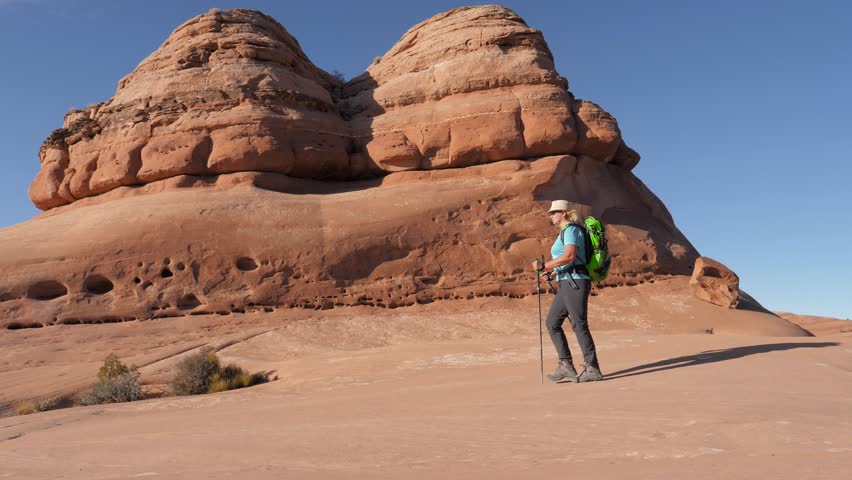 Hiker mature woman with backpack and trekking poles hiking at flat and smooth rock monolith of orange sandstone. Walking background rock formations in Arches national park Utah. Slow motion side view