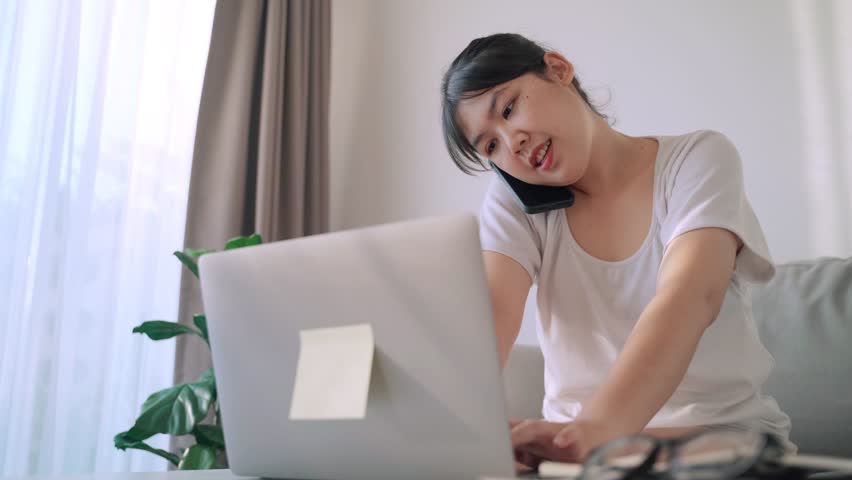 Productive Asian woman on a phone call and typing on a laptop computer keyboard. | Shutterstock HD Video #1101039351