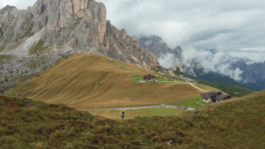 Tourist walks on hill top edge exploring high mountain Giau pass under cloudy sky. Hiker with backpack looks at small village locating in mountain valley