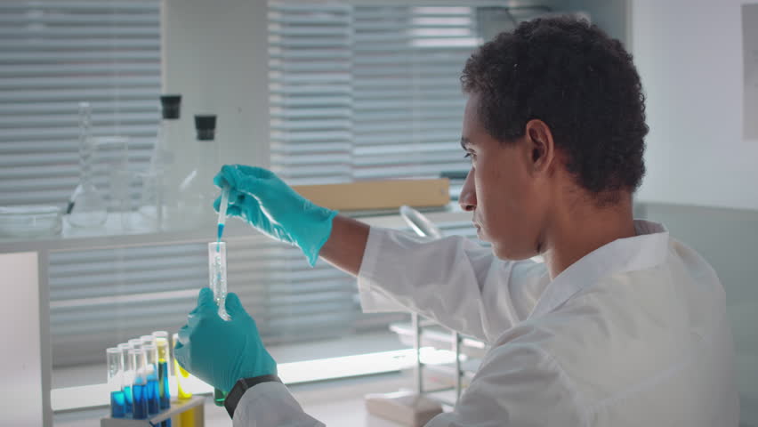 Focused young Biracial male biotechnologist in sterile gloves and white lab coat doing experiment in laboratory, mixing liquids in glass test tube with pipette | Shutterstock HD Video #1101041853