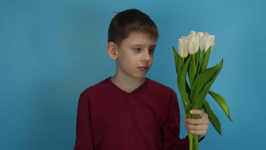 gallant caucasian boy 9 years old with a bouquet of flowers waiting for a late girlfriend on a date, looking at the clock upset and dissatisfied, blue background studio shooting Royalty-Free Stock Footage #1101043039