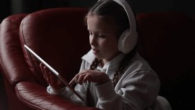 Concentrated teen girl in headphones plays game, communicate on internet social media, listens to music or online studies at home on digital tablet sitting on sofa. Children tech addiction concept.