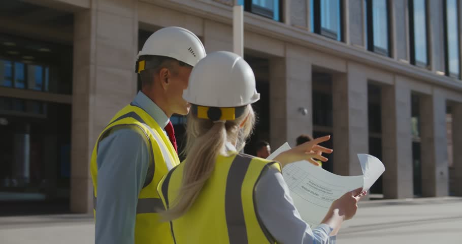 Professional architects in helmet studying blueprint outside modern building. Civil engineers discussing project holding blueprint at construction site Royalty-Free Stock Footage #1101044989