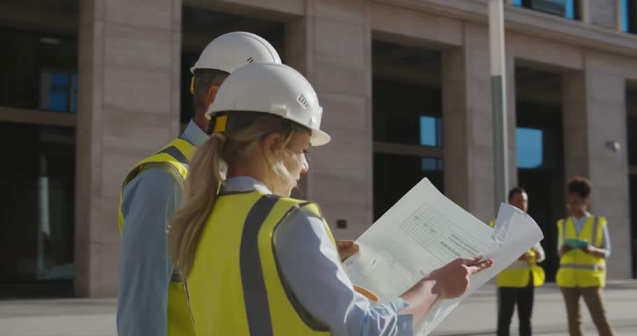 Professional architects in helmet studying blueprint outside modern building. Civil engineers discussing project holding blueprint at construction site