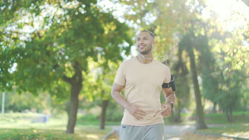 The young runner of mixed race with headphones and a smartphone jogging in an urban city park. A beautiful man in a tracksuit likes to run outdoors under his favorite music. Нealthy lifestyle concept Royalty-Free Stock Footage #1101051353