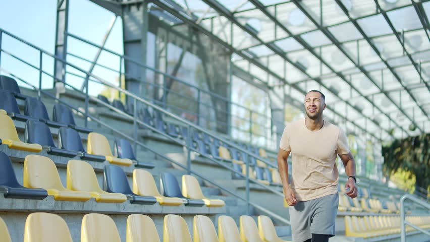 Tired young runner has shortness of breath after running in urban city stadium. Exhausted male of mixed race stands with his hands on his knees, breathes deeply, wipes sweat from his forehead Close up Royalty-Free Stock Footage #1101051377