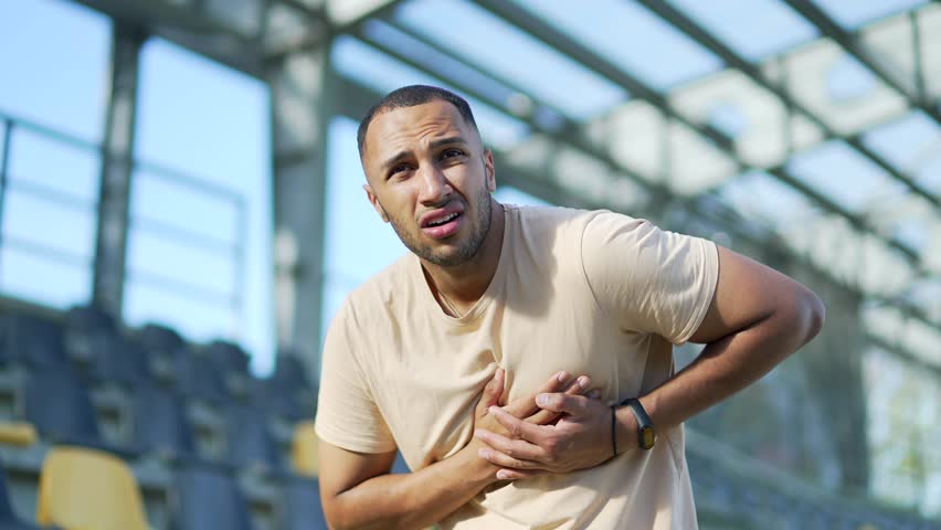 Close up Young athlete survives a heart attack while standing in an urban city stadium. Sad male suffers, feels tension in the chest, holds hands to a sore spot. He needs urgent medical attention | Shutterstock HD Video #1101051379