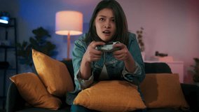 Excited beautiful Asian woman using joystick wireless controller enjoy playing console video game glad to win the match get highest victory sit on sofa at home in RGB living room. Hand held Video shot