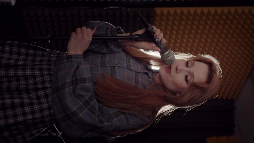 A young woman recording a song in the sound recording studio - singing in the mic and looking in the camera