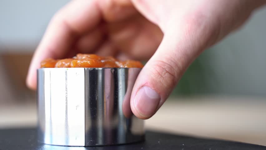 hand taking out metal cast of salmon tartar with avocado and seaweed decorated with sesame seeds over black stone