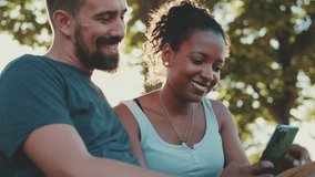 Happy smiling couple watching video on mobile phone while sitting on bench