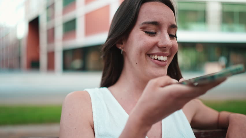 Beautiful woman with freckles and dark loose hair wearing white top sits on bench with phone in her hands. Cute girl sends voice message on mobile phone modern city background | Shutterstock HD Video #1101067347