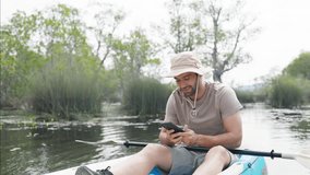 4K Caucasian man using smartphone taking video during kayaking at mangrove forest on summer vacation. Handsome guy enjoy outdoor lifestyle canoeing on lake. Environmental ecotourism travel concept.