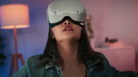 Young Beautiful Asian woman putting on Virtual Reality headset exploring playing VR world game with curious and fun pointing fingers on screen sitting on sofa at home in RGB living room in the evening