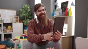 Young redhead man preschool teacher having video call sitting on chair at kindergarten