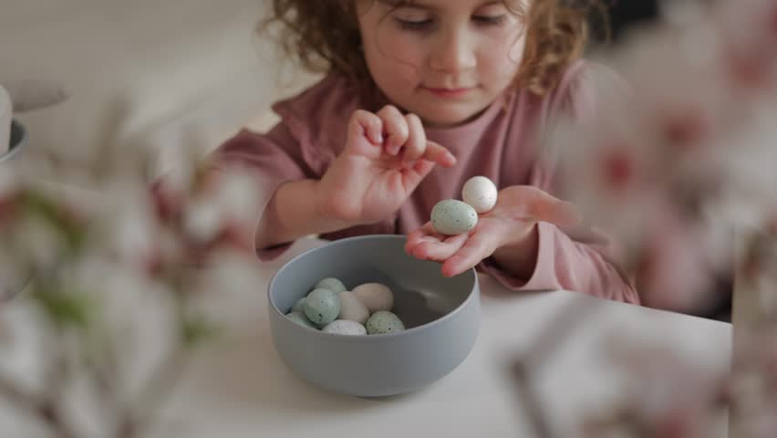 View of hands of pretty girl playing with Easter eggs. She is sitting at a table with spring flowers, almond blossom, looking and touching the eggs celebrating Happy Easter. Royalty-Free Stock Footage #1101071015