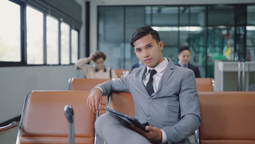 Close up smiling face of young Asian businessman wearing suit sitting at airport terminal looking at camera