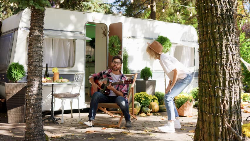 Funny young couple. A guy plays the guitar and a girl dances in front of a travel trailer. Rest outside the city. Happy people enjoy. Weekends in the summer time. Romance in nature.