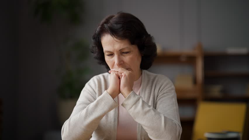 Sad senior woman sits at home and thinks, putting chin on hands crossed in castle. Thinks about important matters and problems. Consider big decision. Beautiful woman sitting on sofa in living room. | Shutterstock HD Video #1101082555