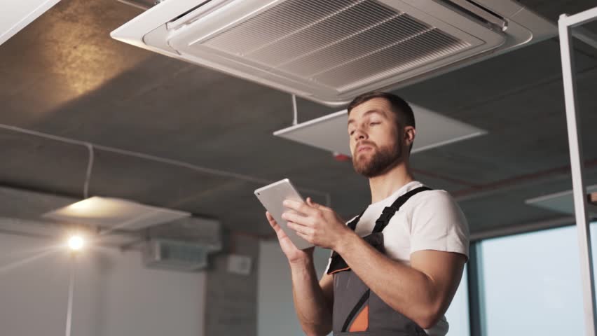 A portrait of an adult man in work uniform with a tablet installing industrial air conditioning and configuring software in a modern office Royalty-Free Stock Footage #1101088311