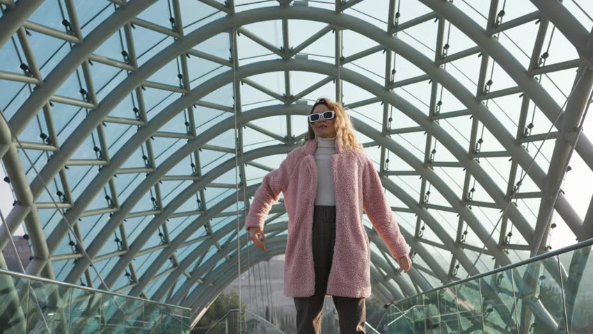 Stylish woman in white glasses and a pink fur coat with flowing hair is twirling on the modern peace bridge in Tbilisi in Georgia. Girl tourist enjoys the journey.