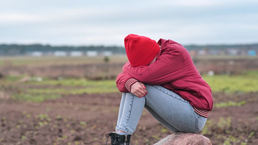 A sad teenage girl sits on a rock in a field and cries.