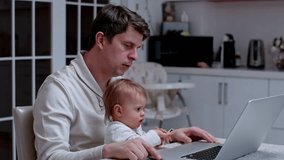 Father working at home with toddler kid, father's day. Man sitting in the kitchen with infant and trying to type on a laptop.