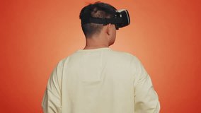 Man using headset helmet to play simulation game app online. Watching virtual reality 3D 360 video content. Guy in VR goggles isolated on studio orange background. Futuristic technology. Rear view