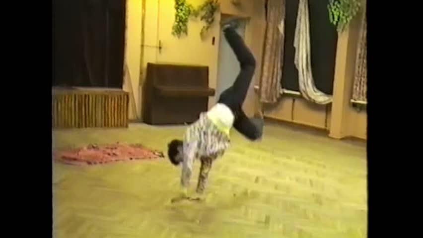 A high school student in the auditorium of the school shows elements of breakdancing, standing on his hands, doing a moonwalk and waves with his hands.