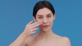 Beautiful woman applying moisturizer to her face. Video of a smiling woman with perfect makeup on a blue background. Beauty concept
