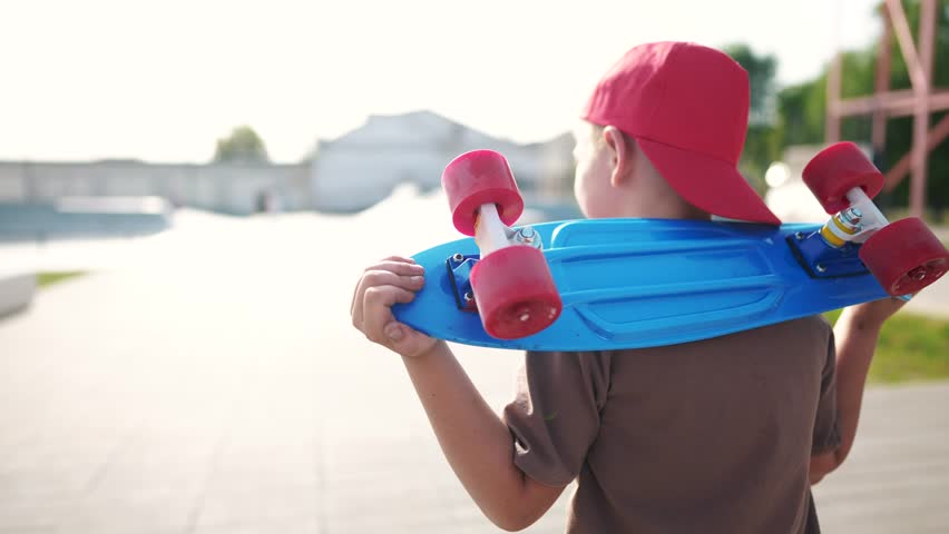 child walk with skateboard. boy in a red cap with a skateboard on the playground portrait. skateboarder child walk outdoors sun glare. kid skateboarder to the skatepark lifestyle